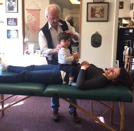Alexander Technique teacher Tommy Thomson performing a hands-on posture session, gently guiding the head of a student lying on a table while a small child rests on the student's stomach. A moment of embodied awareness, family presence, and quiet connection.