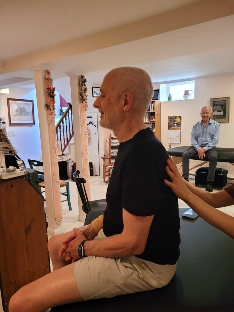 A trainee receives gentle back contact during a seated moment in a Tommy Thompson Alexander Technique class, demonstrating inhibition in movement in daily posture.