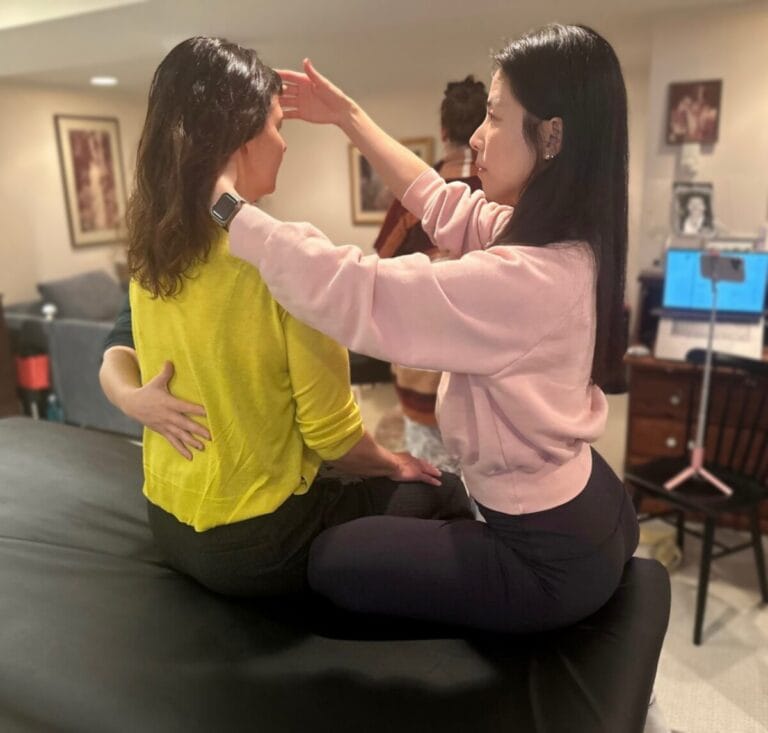 A trainee in a pink sweatshirt gently guides another’s head during a Tommy Thompson-led Alexander Technique class, illustrating inhibition in movement.