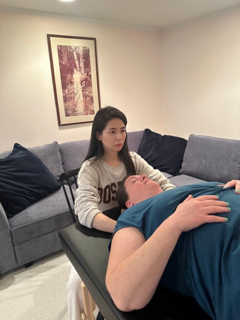 Trainee Ari offering gentle contact to fellow trainee Lili during tablework in Alexander Technique Class 51, exploring presence and relational touch